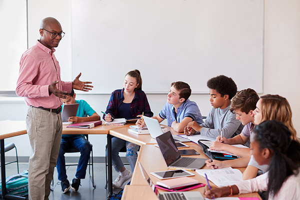 High School Teacher Talking To Pupils Using Digital Devices In Technology Class